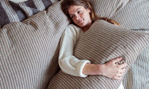 Eine Frau mit langen braunen Haaren liegt auf einem Bett, trägt einen weißen Pullover und schmiegt sich an ein gestreiftes Kopfkissen. Sie schaut nachdenklich, umgeben von San Gabriel Stripe Leinen Bettwäsche von Libeco.