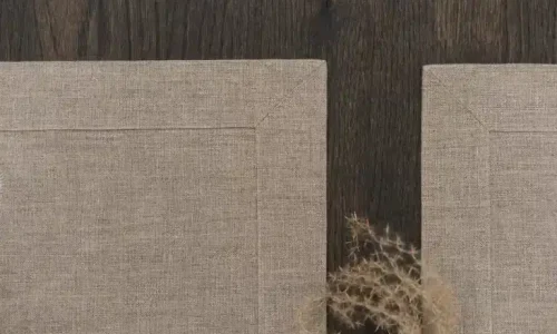 Zwei beigefarbene Stoff-Tischsets liegen auf einer dunklen Holzfläche, wobei sich die Ecken fast berühren. Ein kleiner Zweig mit getrocknetem Gras liegt teilweise auf dem rechten Tischset.