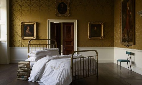 Ein Vintage-Schlafzimmer mit einem Messingbett mit Shanghai Percal 400 TC Bettwäsche von Peter Reed, weißer Bettwäsche, gestapelten Matratzen am Fußende, Goldtapete, vier Porträts, einer Holztür, einem Fenster und einem tealfarbenen Stuhl sorgt für einen eleganten Look.