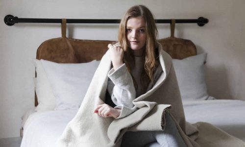 Eine junge Frau mit langen Haaren sitzt auf einem ordentlich gemachten Bett, eingewickelt in eine beigefarbene Decke. Sie trägt bequeme, legere Kleidung und blickt in einem sanft beleuchteten Schlafzimmer direkt in die Kamera.