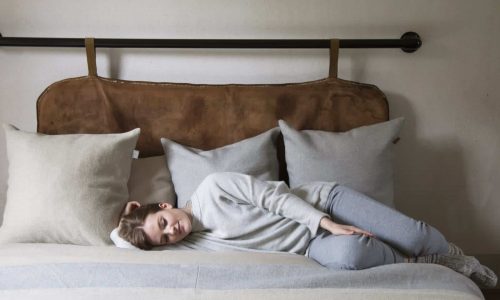 Eine Person mit kurzen Haaren in einem grauen Outfit liegt auf der Seite auf einem Bett mit beigen und grauen Kissen und stützt ihren Kopf auf ein Kissen in einem gemütlichen, minimalistischen Schlafzimmer.