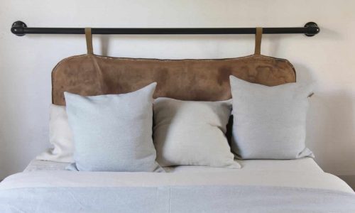 Ein ordentlich gemachtes Bett mit einem braunen Lederkopfteil, drei hellgrauen Kissen und einer weißen Tagesdecke. Eine schwarze Stange mit Lederriemen stützt das Kopfteil vor einer schlichten weißen Wand.