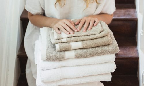 Eine Frau in einem weißen Kleid sitzt auf einer Holztreppe und hält einen Stapel Simi Leinenfrottier fürs Bad Flax von Libeco in der Hand. Ihre Hände ruhen sanft auf der sauber gefalteten weißen und beigen Wäsche.