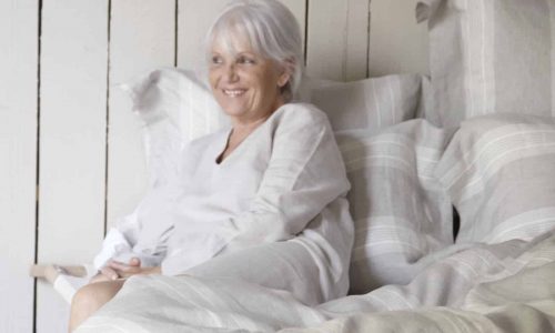 Eine ältere Frau mit grauem Haar und einem hellen Oberteil sitzt auf einem Bett mit gestreifter Bettwäsche, lächelt und entspannt sich vor dem Hintergrund einer Holzwand.