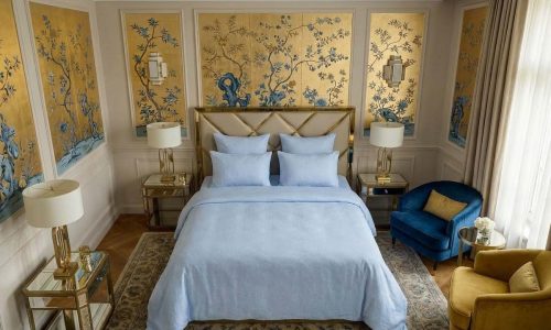 Elegantes Schlafzimmer mit goldener und blauer Blumentapete, einem Bett mit Sol Halbleinen-Bettwäsche von Schlitzer Leinen, verspiegelten Nachttischen, zwei Lampen und einem goldenen Kopfteil. Blaue und gelbe Stühle stehen an einem großen Fenster mit durchsichtigen Vorhängen.