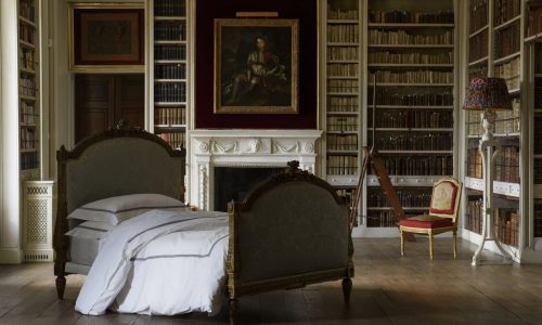 Ein großes Schlafzimmer mit zwei antiken Betten, bezogen mit Stave Percal 400TC Bettwäsche von Peter Reed, weißem Bettzeug, Bücherregalen an den Wänden, einem Kamin mit einem Porträt, einem kleinen roten Stuhl und einer Stehlampe mit einem bunten Schirm.