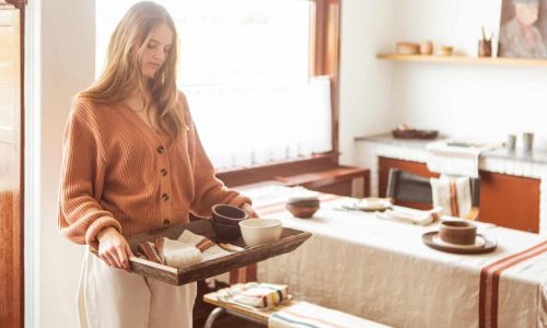 Eine Frau in einem gemütlichen, sonnendurchfluteten Esszimmer trägt ein Holztablett mit Tassen und Schüsseln. Der Tisch ist mit einer gestreiften Tischdecke und einfachem, rustikalem Geschirr gedeckt, was eine warme, einladende Atmosphäre schafft.