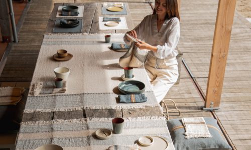 Eine Frau faltet eine Stoffserviette an einem langen, mit Tellern, Tassen und Schüsseln gedeckten Tisch auf einer Holzterrasse. Der Tisch ist mit der belgischen Tischdecke Ioulida reines Leinen von Libeco drapiert, während natürliches Licht einfällt.