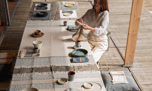 Eine Frau faltet eine Stoffserviette an einem langen, mit Tellern, Tassen und Schüsseln gedeckten Tisch auf einer Holzterrasse. Der Tisch ist mit der belgischen Tischdecke Ioulida reines Leinen von Libeco drapiert, während natürliches Licht einfällt.