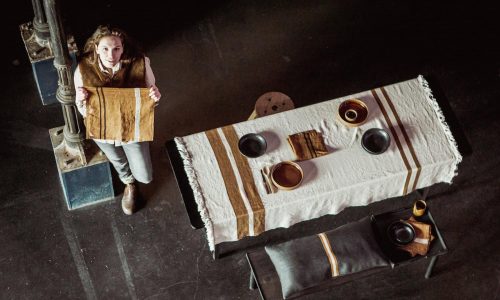 Eine Person steht neben einem Tisch mit Tellern, Schüsseln und Tassen auf einem beigen Tischtuch mit braunen Streifen. Die Person hält ein passendes Tuch und blickt nach oben. Die Szene ist von oben in einem schwach beleuchteten Raum zu sehen.