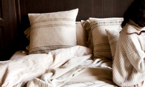 Auf einem gemütlichen Bett liegt der Marokkanische Streifen Leinen-Wolle-Mix von Libeco in beige-weiß gestreift. Auf der rechten Seite sitzt eine Person mit langen dunklen Haaren in einem hellen Strickpullover, der eine warme, entspannte Atmosphäre schafft.