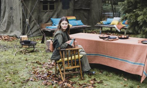 Eine Frau sitzt an einem mit einem gestreiften Tuch bedeckten Tisch im Freien, umgeben von Herbstlaub. Hinter ihr stehen Bänke mit bunten Kissen und ein großes grünes Zelt, das auf ein gemütliches Treffen im Freien oder einen Campingplatz hindeutet.