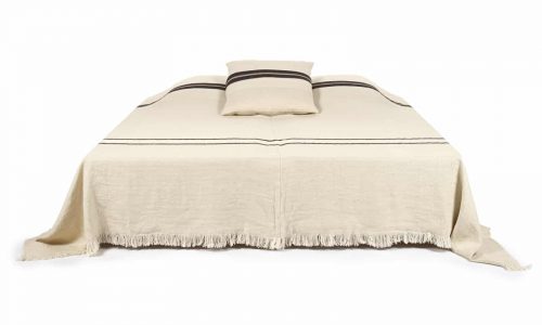 Ein beigefarbenes Bett mit fransenbesetzter Tagesdecke und passendem Kissen, beide mit schwarzen Querstreifen, steht vor einem weißen Hintergrund.