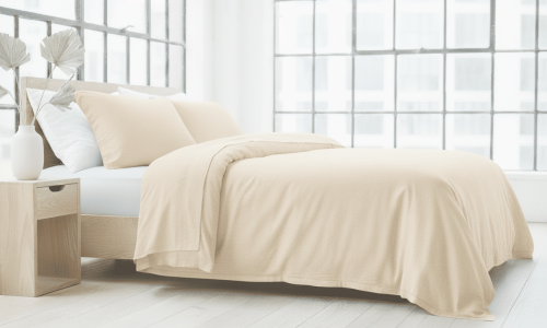 Ein modernes Schlafzimmer mit einem großen Bett, bezogen mit Hanf und Leinen Bettwäsche Oyster von Vieböck 226/02, zwei Kissen, einem hölzernen Nachttisch mit einer weißen Vase und großen Fenstern, die den hellen, minimalistischen Raum mit natürlichem Sonnenlicht füllen.