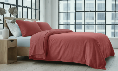 Ein modernes Schlafzimmer mit großen Gitterfenstern, einem Holzbettgestell, Hanf und Leinen Bettwäsche Burgunder von Vieböck 226/30, einem hellen Holznachttisch und einer weißen Vase mit dekorativen Blättern auf einem hellen Holzboden.