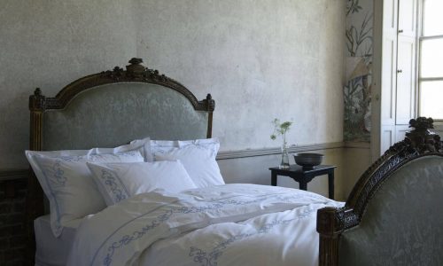 Verziertes Vintage-Doppelbett mit geschnitztem Kopf- und Fußteil, bezogen mit Vienna Percal 400TC Bettwäsche von Peter Reed in Weiß und Blau, in einem sanft beleuchteten Raum mit schlichten Wänden, Fenster und einem schwarzen Beistelltisch mit Vase und Schale.