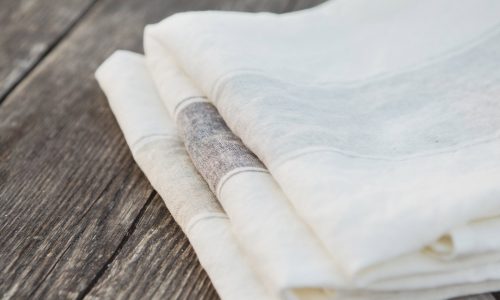 Drei fein säuberlich gefaltete Ajaccio - reines Leinen für die Küche von Libeco Servietten in Weiß und Off-White werden auf einer rustikalen Holzoberfläche präsentiert, die die Texturen von Leinen und Holz hervorhebt.