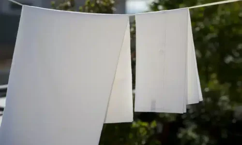 Zwei weiße Hemden, die auf einer Wäscheleine im Freien hängen und im Sonnenlicht trocknen, mit grünen Bäumen und einem unscharfen Gebäude im Hintergrund.