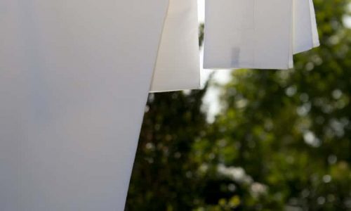 Zwei weiße Kleidungsstücke, darunter ein Hemd, hängen an einer Wäscheleine im Freien mit grünen Bäumen und Sonnenlicht im Hintergrund.