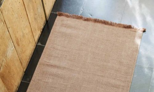 Ein rechteckiger hellbrauner Webteppich mit Fransen an einem Ende liegt auf einem dunklen Kachelboden neben einer Holzwand.