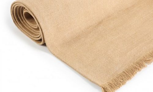 Eine Nahaufnahme des Cambridge Teppichs Wheat reines Leinen von Libeco, aufgerollt mit Fransen an einem Ende, gezeigt vor einem weißen Hintergrund.