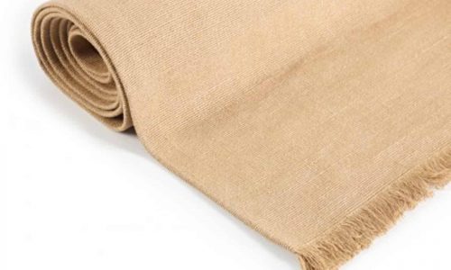 Eine Nahaufnahme des Cambridge Teppichs Wheat reines Leinen von Libeco, aufgerollt mit Fransen an einem Ende, gezeigt vor einem weißen Hintergrund.