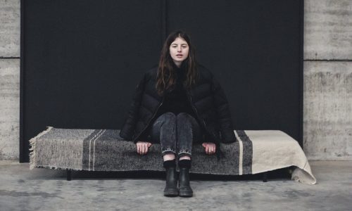 Eine Frau mit langen braunen Haaren sitzt auf einer mit einer gestreiften Decke bedeckten Bank. Sie trägt eine schwarze Pufferjacke, schwarze Jeans und schwarze Stiefel vor einem modernen Hintergrund aus Beton und einer schwarzen Wand.