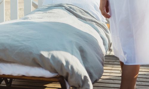 Eine weiß gekleidete Person steht neben einem Outdoor-Bett in Heritage Ash Bio-Leinen Bettwäsche von Libeco, das auf einer Holzterrasse mit einem weißen Zaun im Hintergrund steht.