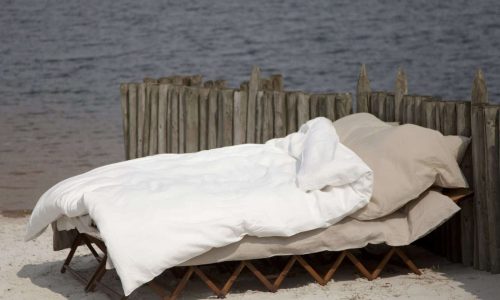 Ein Bett mit Heritage Flax Bio-Leinen Bettwäsche von Libeco liegt an einem Sandstrand in der Nähe eines Holzzauns, mit ruhigem Wasser im Hintergrund.