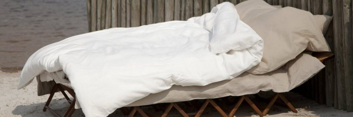 Ein gemütliches Outdoor-Bett mit beigen Laken und Kissen, bedeckt mit einer flauschigen weißen Decke, ruht auf einem klappbaren Holzrahmen in der Nähe eines rustikalen Holzzauns auf sandigem Boden - perfekt zum Entspannen und Staunen: Wie pflege ich Leinenbettwäsche richtig?.