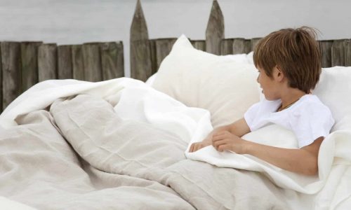 Ein Junge in einem weißen Hemd sitzt auf einem Outdoor-Bett aus Heritage Oyster Bio-Leinen Bettwäsche Libeco und blickt nach rechts. Das Bett befindet sich in der Nähe eines Holzzauns mit ruhigem Wasser im Hintergrund.