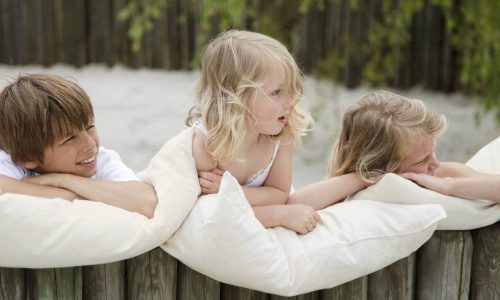 Drei kleine Kinder lehnen an einem mit weißen Kissen bedeckten Holzzaun und schauen zur Seite. Sie wirken entspannt und glücklich im Freien, mit Sand und Bäumen im Hintergrund.
