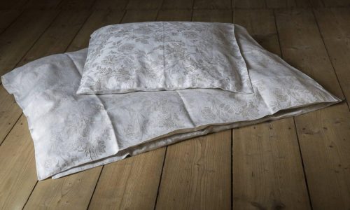 Ein gemustertes weißes Kissen und eine weiße Decke liegen auf einem Holzboden, wobei das sanfte natürliche Licht die Bettwäsche beleuchtet und die Struktur der Holzdielen hervorhebt.