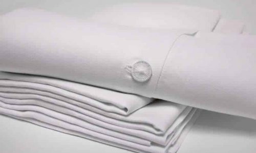 Ein ordentlich gefalteter Stapel Weiße Leinen Bettwäsche mit Hohlsaum von Hoffmann, mit einer dekorativen Knopfverzierung auf einem Stück, ruht auf einer schlichten weißen Oberfläche.