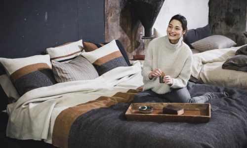 Eine Frau in einem weißen Rollkragenpullover sitzt auf einem Bett mit Nash Leinen-Wolle-Mix von Libeco und hält lächelnd eine Tasse in der Hand. Ein Holztablett mit Snacks und einer weiteren Tasse trägt zur gemütlichen, rustikalen Atmosphäre des Bettes bei.