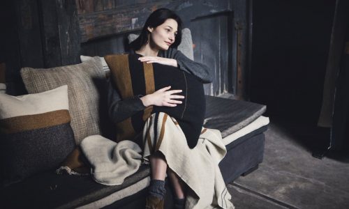 Eine Frau sitzt auf einem Sofa in einem schwach beleuchteten Raum und schmiegt sich an ein Oscar Leinen-Wolle-Mix von Libeco-Kissen. Eingehüllt in eine Decke mit kuscheligen Socken und Stiefeln, blickt sie nachdenklich in die Ferne.