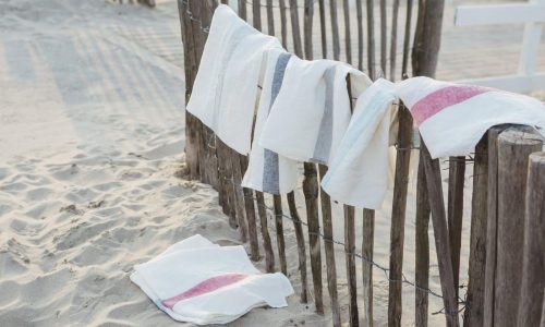 Weiße Handtücher mit farbigen Streifen, darunter die Outlast und Maize Kissen von HEFEL, hängen an einem rustikalen Holzzaun am Strand, wobei ein Handtuch im Sand liegt - so entsteht eine entspannte Küstenatmosphäre.
