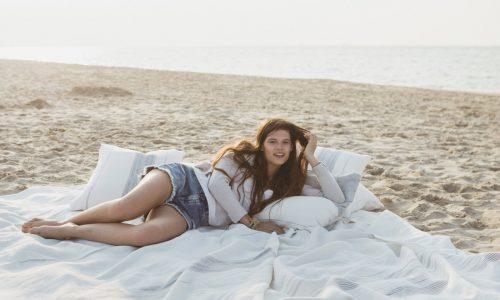 Eine junge Frau mit langen Haaren entspannt sich an einem Sandstrand in der Nähe des Meeres, liegt auf dem Propriano Leinen Coverlet und Kissen von Libeco, ruht ihren Kopf aus und blickt mit ausgestreckten Beinen in die Kamera.