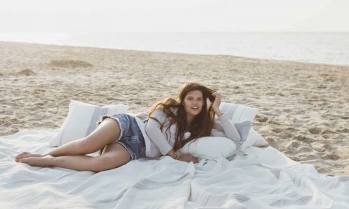 Eine junge Frau mit langen Haaren entspannt sich an einem Sandstrand in der Nähe des Meeres, liegt auf dem Propriano Leinen Coverlet und Kissen von Libeco, ruht ihren Kopf aus und blickt mit ausgestreckten Beinen in die Kamera.