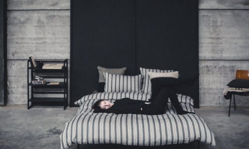 Eine schwarz gekleidete Person liegt auf einem gestreiften Bett aus The Tack Stripe Leinen Bettwäsche von Libeco in einem Raum im Industriestil mit Betonwänden, minimalistischen Möbeln und einem Bücherregal.